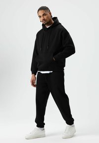 Zwarte hoodie met trekkoorden en een voorzak, gecombineerd met zwarte taps toelopende sweatpants en witte sneakers, tegen een effen witte achtergrond.