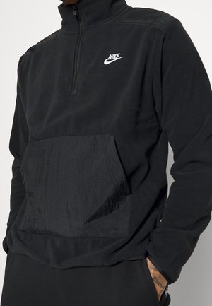Schwarzer Fleece-Pullover mit hohem Kragen, halbem Reißverschluss vorne und einer strukturierten Brusttasche; mit einem weißen Nike-Logo auf der linken Seite.