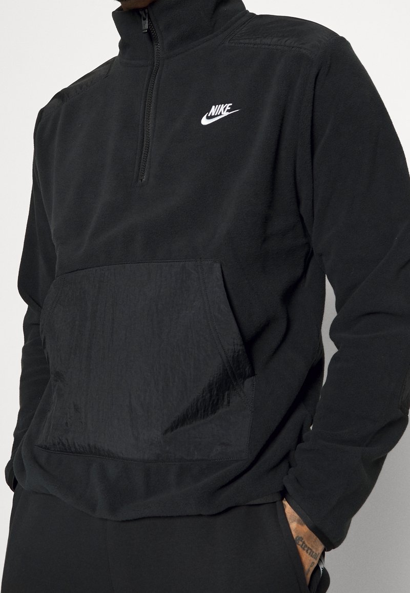 Schwarzer Fleece-Pullover mit hohem Kragen, halbem Reißverschluss vorne und einer strukturierten Brusttasche; mit einem weißen Nike-Logo auf der linken Seite.