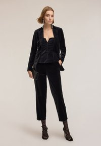 Garnitur z czarnego aksamitu z topem w kształcie litery V o strukturze, dopasowanymi dopasowanymi spodniami oraz czarnymi koronkowymi pończochami w panterkę.