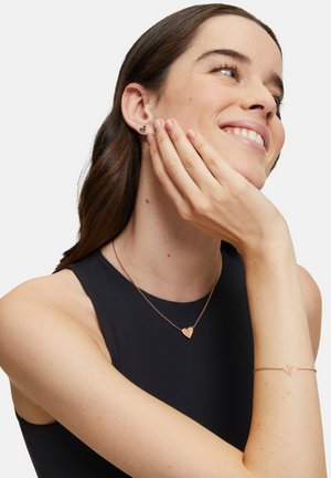 Collier et bracelet en or rose en forme de cœur, avec un design texturé, associés à des boucles d'oreilles puces dans un style similaire. Haut sans manches noir.