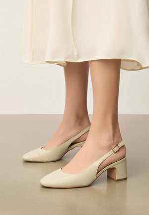 Pieds chaussés de sandales à talon bas carré couleur crème, associées à une robe midi légère et fluide.