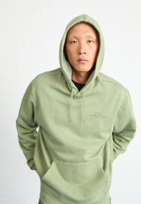 Πράσινο φούτερ με μαλακό ύφασμα, διαθέτει καγκουρό τσέπη, κουκούλα με κορδόνι και μικρό λογότυπο στο στήθος. Κανονική εφαρμογή.