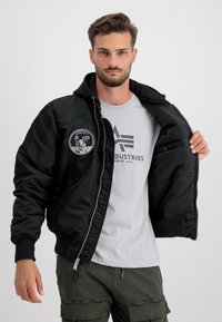 Giacca bomber nera con chiusura a zip e polsini a costine, con un patch ricamato dell'Apollo 11 sul lato sinistro del petto. Indossata sopra una maglietta grigia.