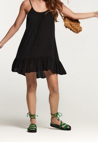 Černé bezramínkové šaty s rozšířeným lemem, z lehkého materiálu. Modelka má na sobě zelené sandály na řemínky a drží texturovanou hnědou kabelku.