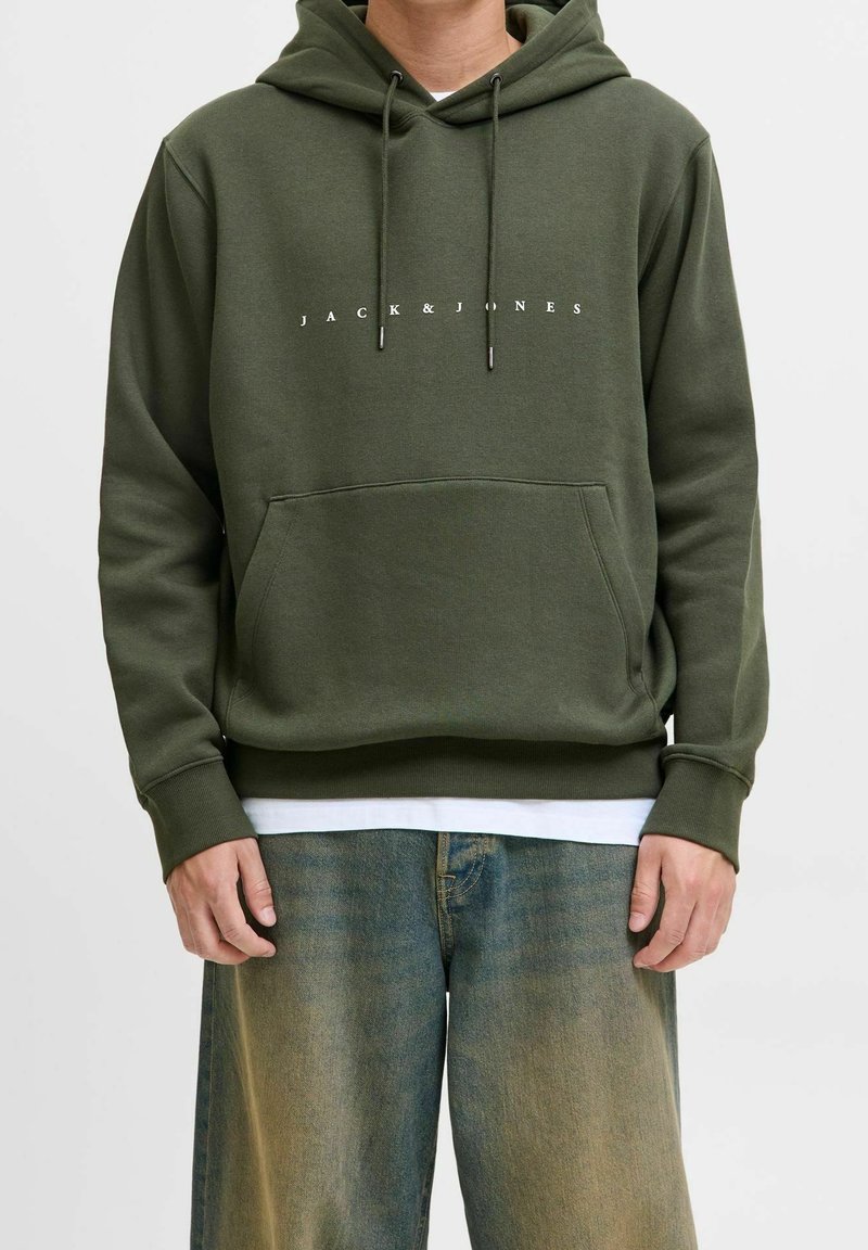 Personne portant un sweat à capuche vert foncé avec le texte "JACK & JONES" et un jean bleu délavé, debout devant un fond clair uni.