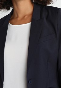 Námořnický modrý blazer s hladkou texturou, zapínání na jeden knoflík a klopovým límcem. Nosený přes bílé blouse s jemným leskem.