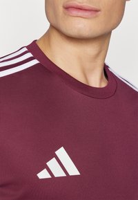 Bordeaux sportshirt met ronde hals, voorzien van witte drie-strepen accenten op de schouders en een wit logo op de borst. Fijne textuur.