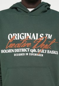 Σκούρο πράσινο hoodie με λευκό και πορτοκαλί κείμενο: "ORIGINALS", "Creative Dept." και "Holmen District cph. Daily Basics", με μπροστινή τσέπη.