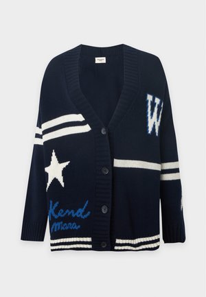 Mujer joven que lleva un cárdigan oversize azul oscuro con estrellas y la letra W, camisa azul, vaqueros de pierna ancha y zapatos marrones puntiagudos.