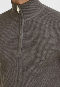 Grauer Reißverschluss-Pullover mit hohem gerippten Kragen und strukturiertem, gemustertem Stoff. Enthält ein goldfarbenes Reißverschlussdetail am Hals.