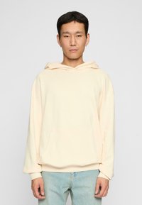 Cream-colored hoodie made of soft material with a front pocket, relaxed fit, and a drawstring hood, paired with light blue jeans.