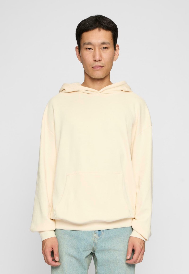 Cream-colored hoodie made of soft material with a front pocket, relaxed fit, and a drawstring hood, paired with light blue jeans.