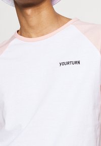 Bílá bavlněná tričko s růžovými raglánovými rukávy, na prsou je černě vyšité heslo "YOURTURN". Hladká textura.