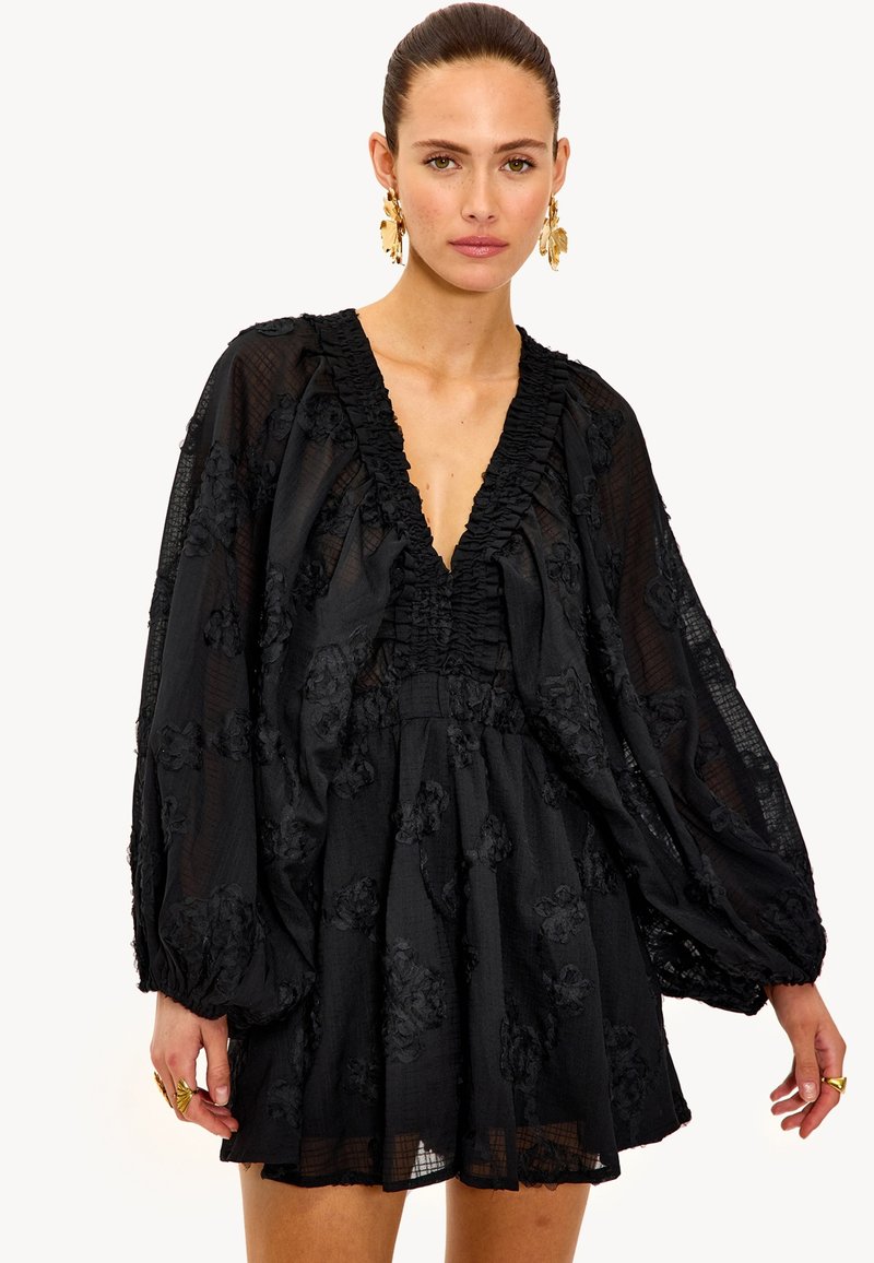 Vestido mini negro con escote en V profundo, tejido texturizado, cuello con volantes y bordado floral. Mangas largas abullonadas y un corte holgado.