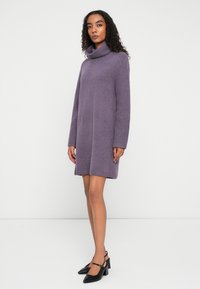 Frau mit lockigem Haar, die ein langärmliges, lavendelfarbenes, gestricktes Rollkragenkleid und schwarze High Heels trägt und vor einem schlichten weißen Hintergrund steht.