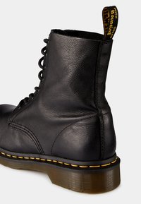 Svart lær snøres boot til anklene med teksturert overflate, gul søm og svart hæl. Har en løftet hanke med "MED DEMPENDE SÅLE."