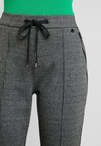 Pantalons à carreaux gris avec une taille élastique à cordon, poches latérales et accents noirs le long des coutures, mettant en valeur un tissu texturé.