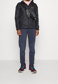 Muž oblečený v černé bundě Columbia, tmavě modrých kalhotách, vínových turistických botách s tyrkysovými tkaničkami a šedé kabelce přes rameno na bílém pozadí.