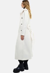 Manteau blanc long en tissu épais, avec de grands boutons, une taille ceinturée et une silhouette droite. Bottes noires épaisses visibles en bas.