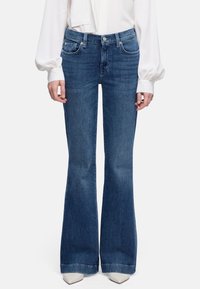 Vrouw draagt donkerblauwe, high-waist wijde jeans, witte blouse met pofmouwen en witte schoenen met puntige neus, staand tegen een witte achtergrond.