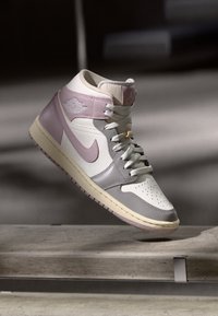 Valge, heleda roosa ja halli värvusega kõrge varbaga tennisekets, millel on laced, keskosa ja Nike logo, näib hõljuvat betoonpinna kohal.