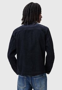Jachetă din cord  de culoare bleumarin închis, cu mâneci lungi și un design curat pe spate. Are o croială slim și cusături structurate. Asortată cu blugi din denim albastru.