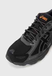 Sapatilha de corrida ASICS preta e cinza com tecido Gore-Tex e atacadores cinza escuro sobre um fundo branco, mostrando detalhes frontais e laterais.