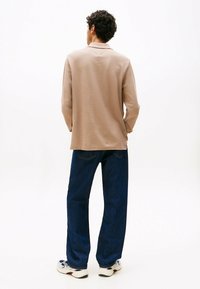Beige Langarmhemd mit Kragen, aus strukturiertem Stoff, kombiniert mit locker sitzenden dunkelblauen Jeans und weißen Turnschuhen.