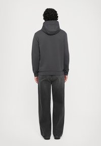 Man with curly hair wearing a dark gray hoodie and loose black jeans, standing facing a plain white background.