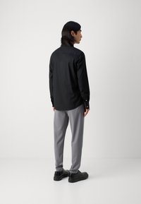Schwarzes Langarmshirt mit Knopfmanschetten; graue Nadelstreifenhose; glattes Material; schmale Passform; getragen mit schwarzen Schuhen. Rückansicht.