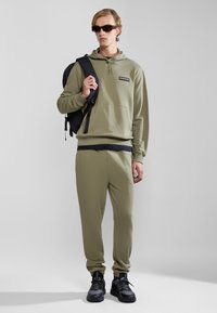 Olive green hoodie with a front pocket, paired with matching joggers. Black sneakers and a black backpack complete the look. Textured fabric.