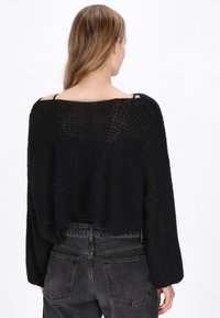 Pulover negru scurt, cu un design texturat, mâneci largi și un spate deschis care evidențiază bretele subțiri. Purtați-l cu blugi gri închis.