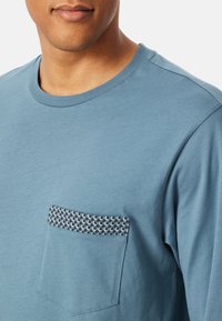 T-shirt en coton bleu avec une poche à motif tissé noir et blanc, encolure ronde et manches courtes. Texture lisse.
