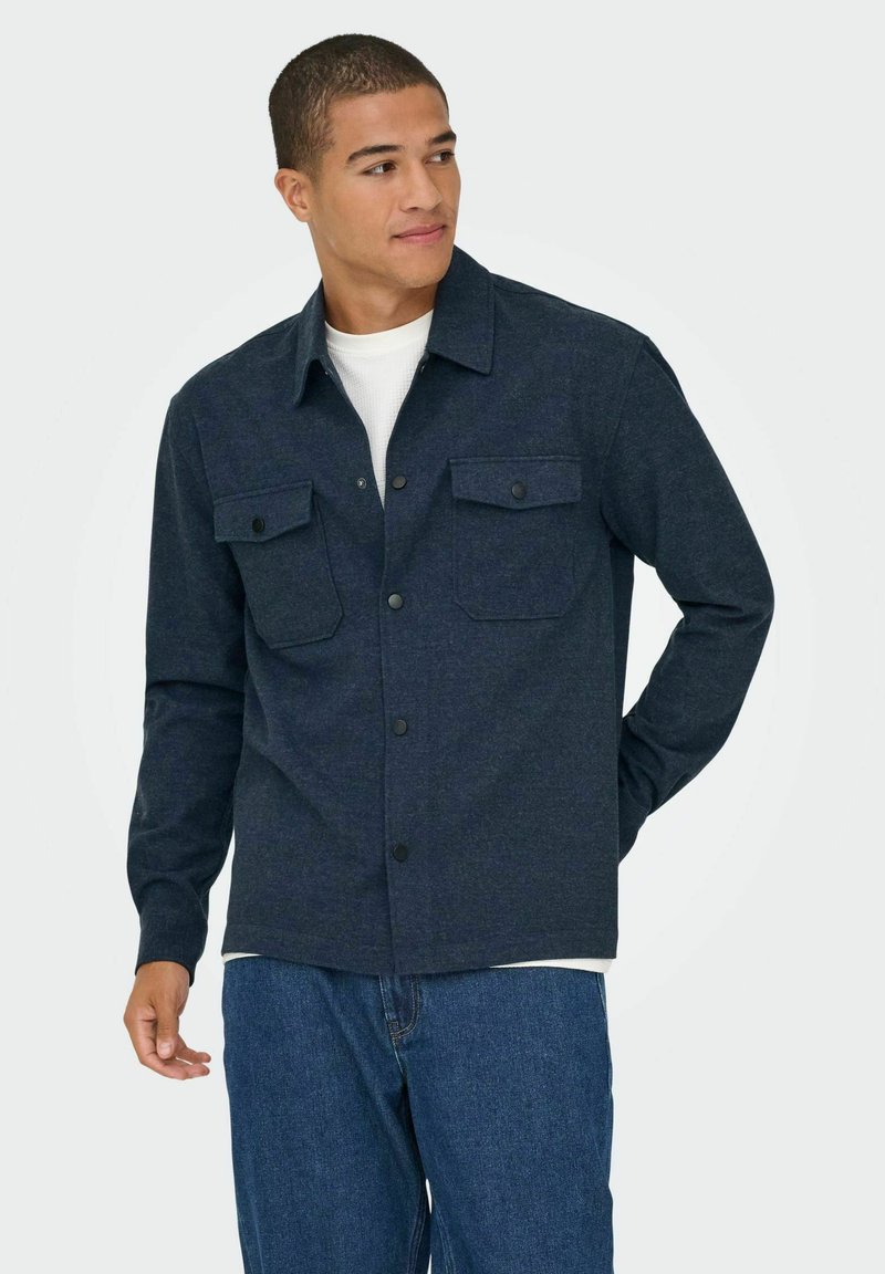 Camisa de botones azul marino confeccionada en tela texturizada, con dos bolsillos en el pecho con solapas, cuello con solapa y mangas largas.