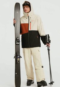 Tenue de ski composée d'une veste beige, noire et orange, de pantalons beiges et de bottes de ski noires ; tenant des skis et des bâtons. Matériaux texturés visibles.