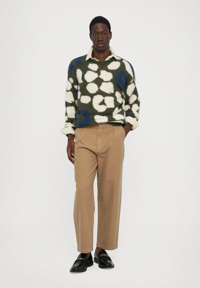 Jersey grueso en verde con patrones blancos y azules, combinado con pantalones beige y mocasines negros. El jersey tiene un corte suelto y puños doblados.