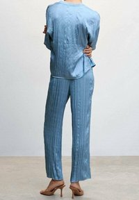 Completo in satin blu chiaro, texturizzato, composto da una blusa ampia e pantaloni dritti. Accentuato da righe verticali e una leggera lucentezza. Tacchi chiusi sul retro.