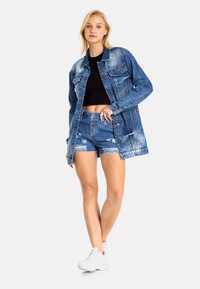 Leren spijkerjack in lichtblauw met een vervaagd uiterlijk, gecombineerd met versleten blauwe shorts met knoopsluiting en rafelige randen. Witte sneakers.