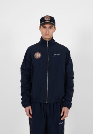 Man draagt een marineblauw trainingspak met een jasje met rits en bijpassende pet, beide met ronde rood-witte patches en witte tekstlogo's.