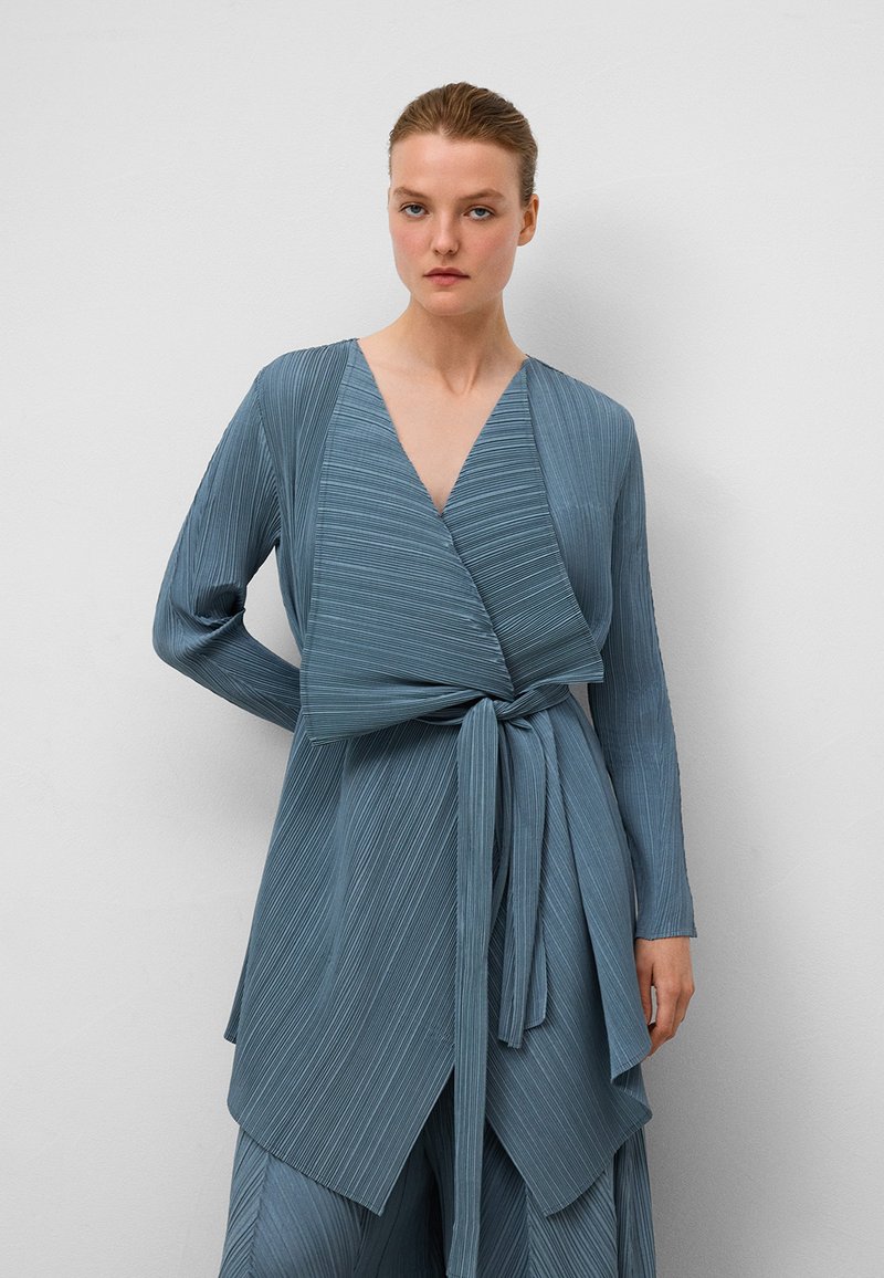 Niebieska, plisowana bluzka z długimi rękawami i wiązaniem z przodu. Posiada dekolt w kształcie litery V oraz warstwowy dół, wykonana z lekkiej, teksturowanej tkaniny.