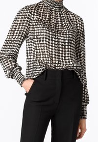 Blusa a motivi bianco e nero con collo alto e maniche lunghe, caratterizzata da un design arricciato e tessuto testurizzato. Abbinata a pantaloni neri.