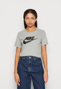 Pilkas medvilninis marškinėlis su juodu Nike logotipu centre, turintis klasikinį apvalų kaklą ir trumpas rankoves, derinamas su tamsiai mėlynomis džinsomis.