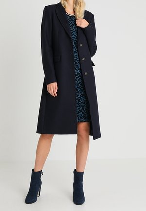 Femme portant un long manteau bleu marine sur une robe bleue à motifs et des bottines bleu marine assorties, debout devant un fond clair uni.