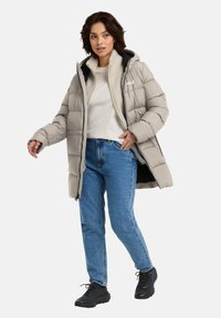 Lichtbeige pufferjas met een capuchon, vooraan met rits en een zachte fleece binnenkant, gedragen met blauwe jeans en zwarte buitenschoenen.