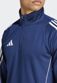 Blaue sportliche Zip-Jacke mit hohem Kragen, ausgestattet mit weißen Schulterstreifen und einem kleinen weißen Logo auf der Brust, mit glatter Textur.