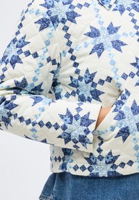 Πουπουλένο μπουφάν με σχέδιο πατς με μπλε και λευκά αστέρια, το χέρι στην πλαϊνή τσέπη, συνδυασμένο με μπλε τζιν παντελόνι.