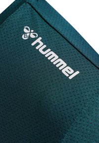 Donkerblauwgroene gaasstof met een gestructureerd oppervlak; beschikt over een wit "hummel" logo en een bij-embleem. Strakke, atletische uitstraling.