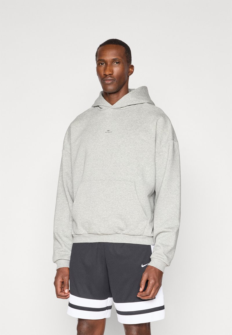 Grey hooded sweatshirt in soft fabric, featuring a front pocket and a small logo at the center. Paired with black shorts with white stripes.