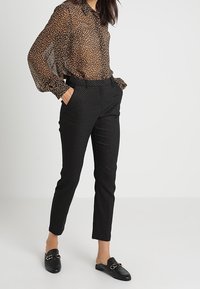 Brązowa koszula z przezroczystym nadrukiem w panterkę, czarne dopasowane spodnie w wzory oraz czarne loafersy z detalami w złotym kolorze. Gładkie tekstury na całej długości.